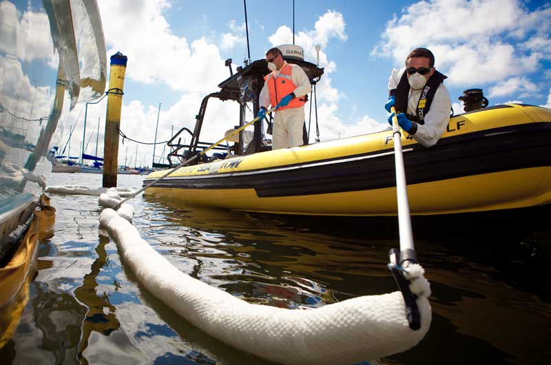 Two workers deploying an oil containment boom from a boat on water.